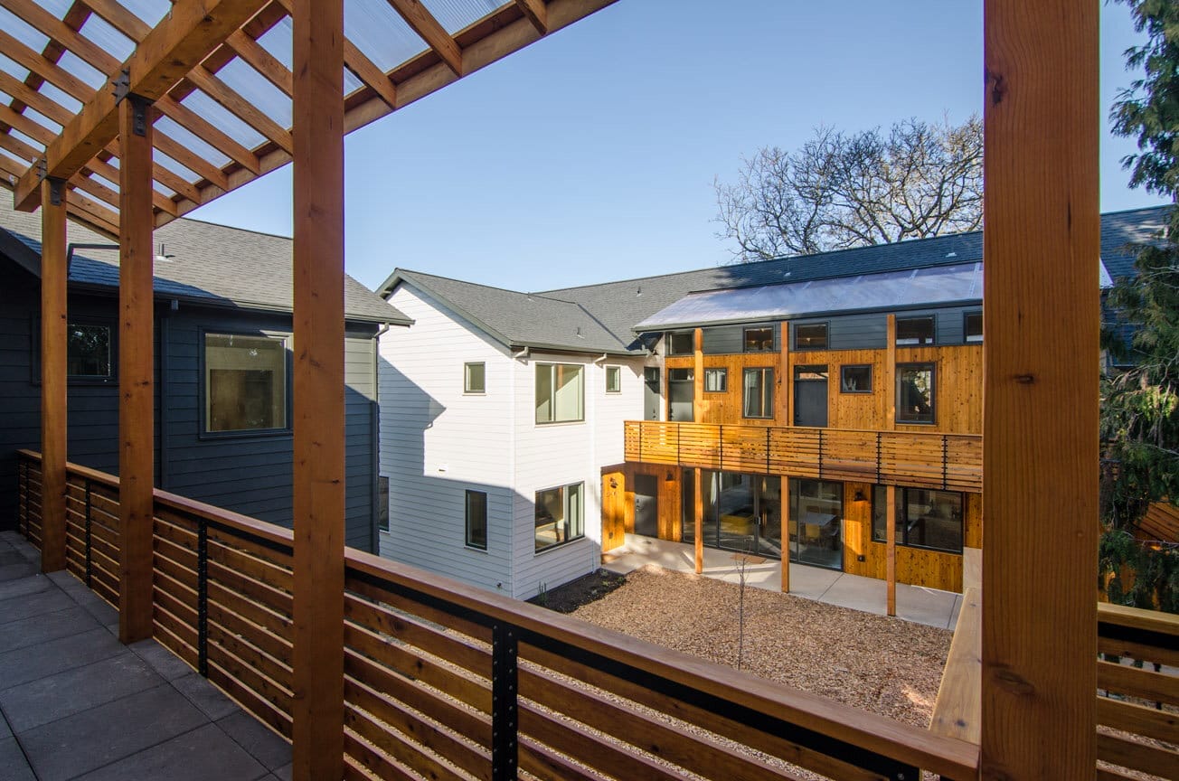 A wooden railing with horizontal slats in the foreground frames a courtyard, a distant building, and blue sky. One section of the building has white siding, and one section has wooden siding.